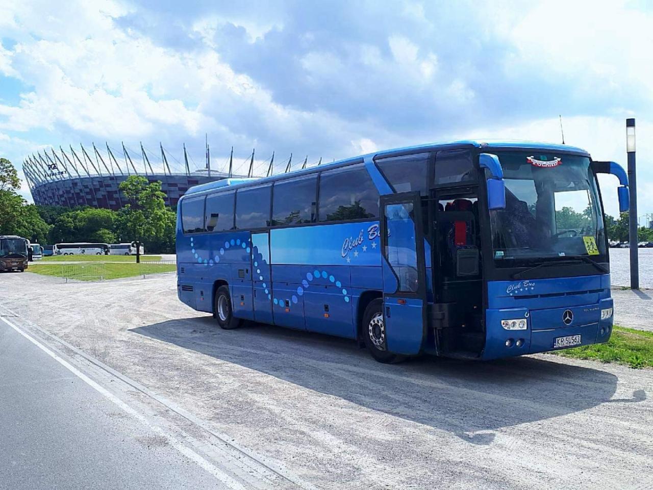 Niebieski autokar turystyczny z otwartymi drzwiami na tle Stadionu Narodowego w Warszawie, z widocznym logo firmy Club Bus i polską flagą na przedniej szybie.