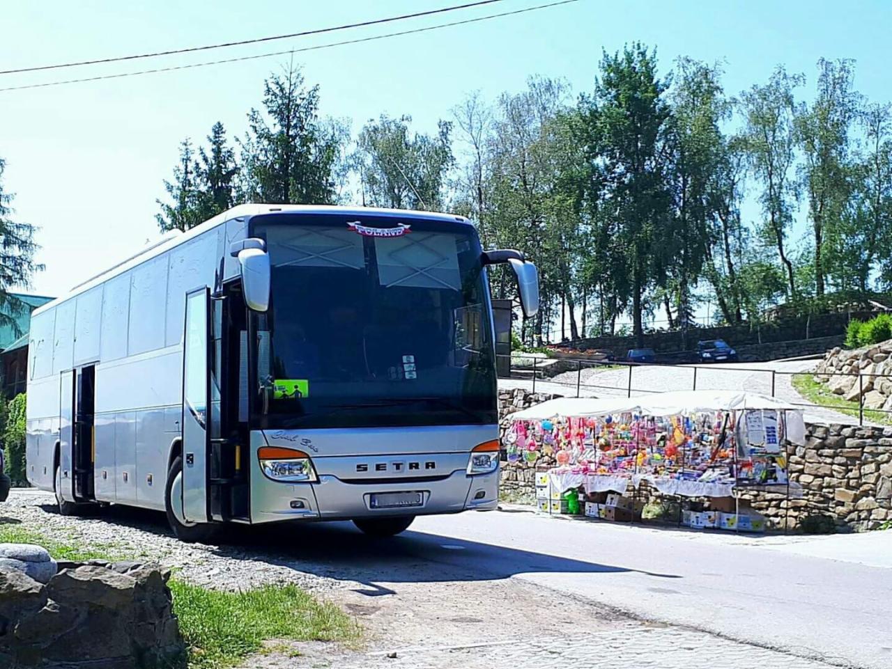 Srebrny autokar Setra z otwartymi drzwiami na tle krajobrazu z drzewami i straganem z pamiątkami, w słoneczny dzień.