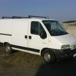 Biały samochód dostawczy Citroen Jumper z relingami dachowymi, zaparkowany na ziemi z widocznym ogrodzeniem w tle.