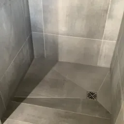 Szara kabina prysznicowa z odpływem liniowym i płytkami imitującymi beton, ułożonymi ze spadkiem w kierunku kratki odpływowej.