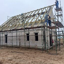 JERZY RODAK - Budowa domu z bloczków: murarze na rusztowaniu montują drewnianą konstrukcję dachu. Widoczne ściany z otworami okiennymi i błotniste podłoże.
