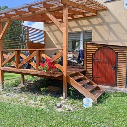 JERZY RODAK - Drewniana altana z przezroczystym zadaszeniem, schodami i czerwoną doniczką z kwiatami. Obok budynku widoczna drewniana budka z czerwonymi drzwiami.