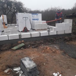 JERZY RODAK - Wznoszenie ścian z białych bloczków SOLBET na fundamencie. Murarz z czerwoną pacą wyrównuje bloczki. Palety z bloczkami i wózek paletowy w tle. Grunt wokół.