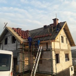 JERZY RODAK - Budowa domu z białego pustaka: dekarze na rusztowaniu układają czerwoną dachówkę. Widoczny komin i fragment białego busa. Częściowo pokryty dach.