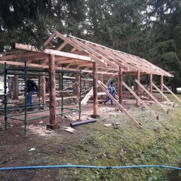 JERZY RODAK - Budowa drewnianej konstrukcji z bali na zewnątrz, widoczne rusztowania i pracownicy w trakcie montażu dachu. W tle drzewa.