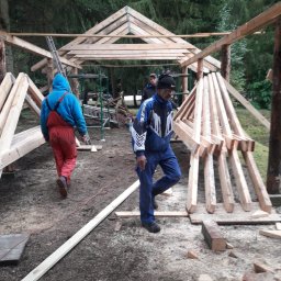 JERZY RODAK - Budowa drewnianej konstrukcji dachu w lesie. Widoczni pracownicy w kombinezonach, drewniane elementy konstrukcyjne i leśne otoczenie. Prace w toku.