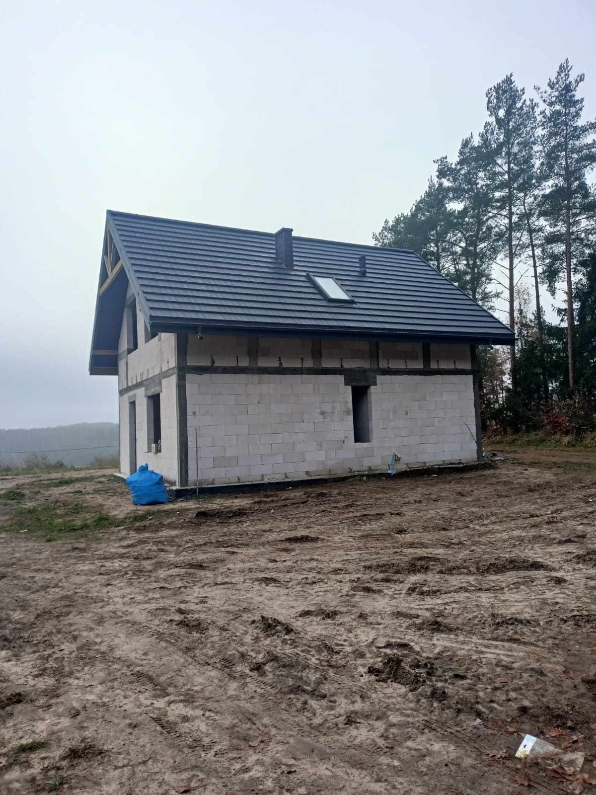 Surowy dom murowany z ciemnym dachem i oknem dachowym, w trakcie budowy. Widoczne białe bloczki, brak elewacji, teren wokół nierówny, zalesione tło.