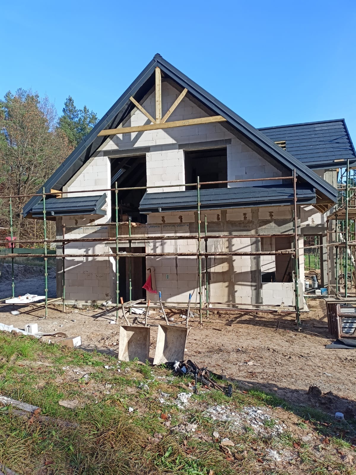 Surowy dom z białego pustaka z czarnym dachem w trakcie budowy, otoczony rusztowaniami i narzędziami na tle lasu i błękitnego nieba.