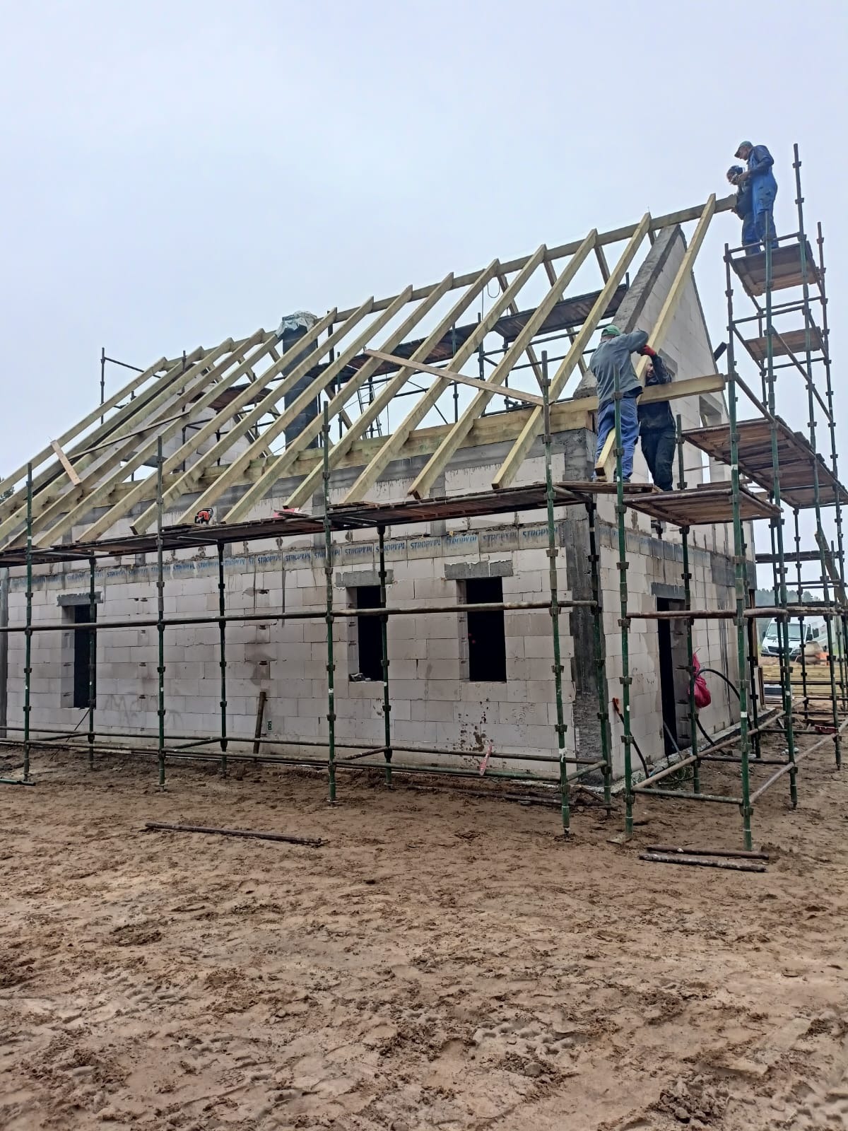 Budowa domu z bloczków: murarze na rusztowaniu montują drewnianą konstrukcję dachu. Widoczne ściany z otworami okiennymi i błotniste podłoże.