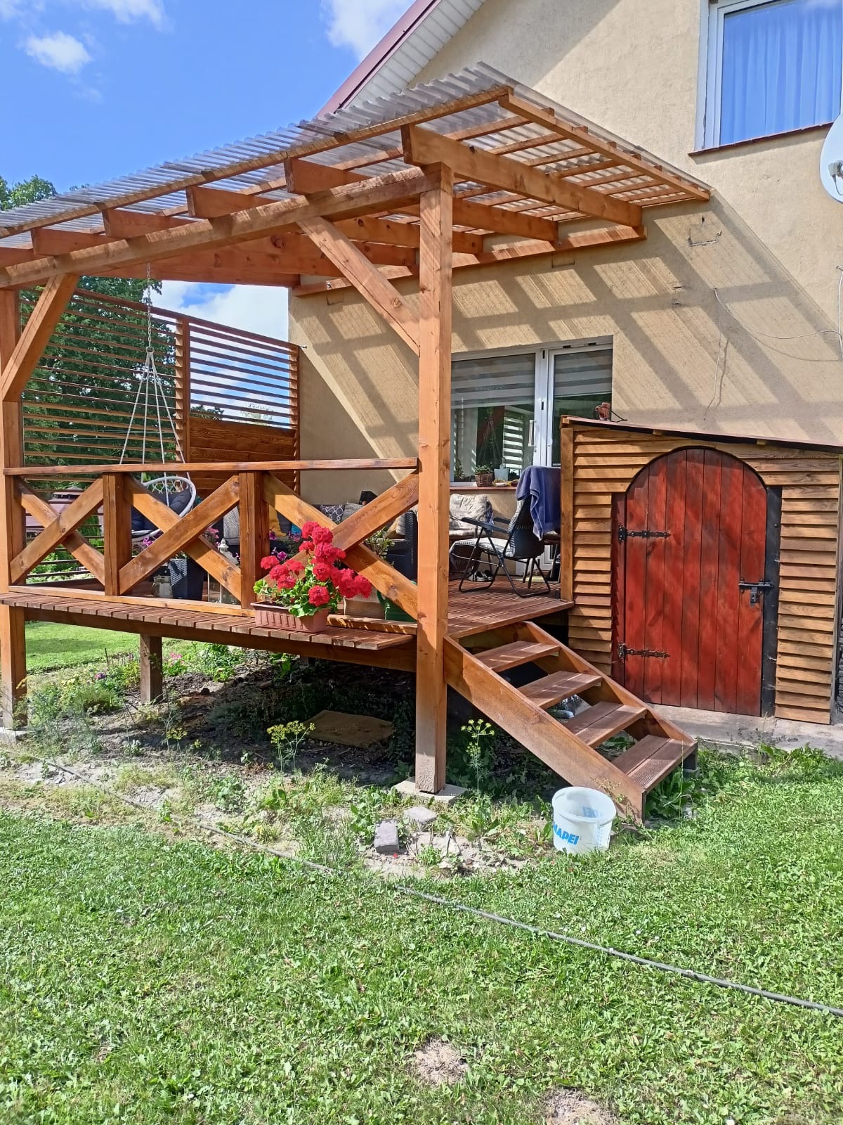 Drewniana altana z przezroczystym zadaszeniem, schodami i czerwoną doniczką z kwiatami. Obok budynku widoczna drewniana budka z czerwonymi drzwiami.