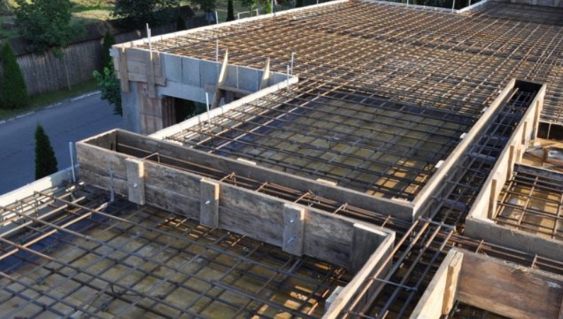 Zbrojenie stropu budynku w trakcie budowy, widoczne drewniane szalunki i siatka z prętów stalowych. Ujęcie z góry, w tle zieleń i fragment drogi.