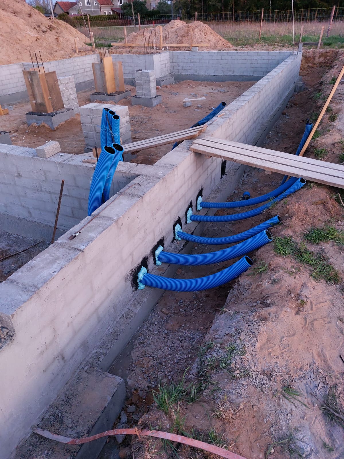 Fundamenty z przepustami na instalacje, zabezpieczone pianką montażową. Widoczne niebieskie rury karbowane. W tle hałda ziemi i budynek w trakcie budowy.