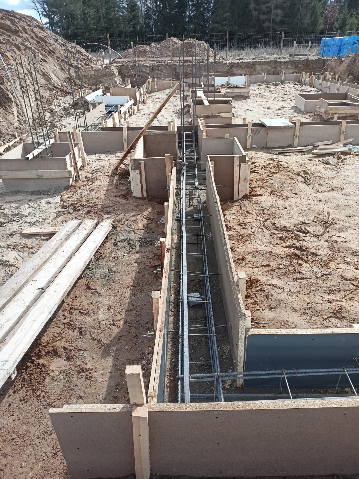 Zbrojenie i szalunki pod fundamenty w Dobre Miasto. Widoczne pręty zbrojeniowe w drewnianych formach, przygotowane do zalania betonem. Grunt pod budowę.