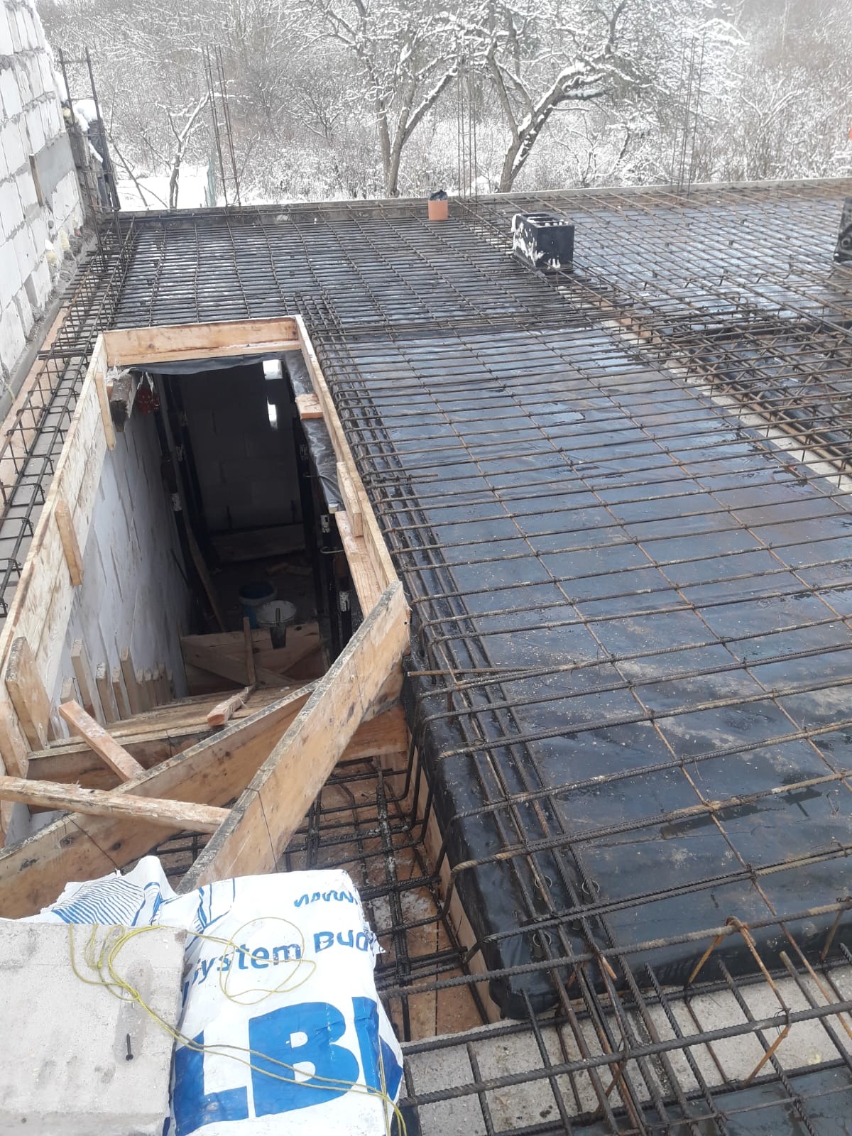 Zbrojenie stropu z otworem na schody, widok z góry. Na pierwszym planie worek z zaprawą budowlaną. W tle ośnieżone drzewa.