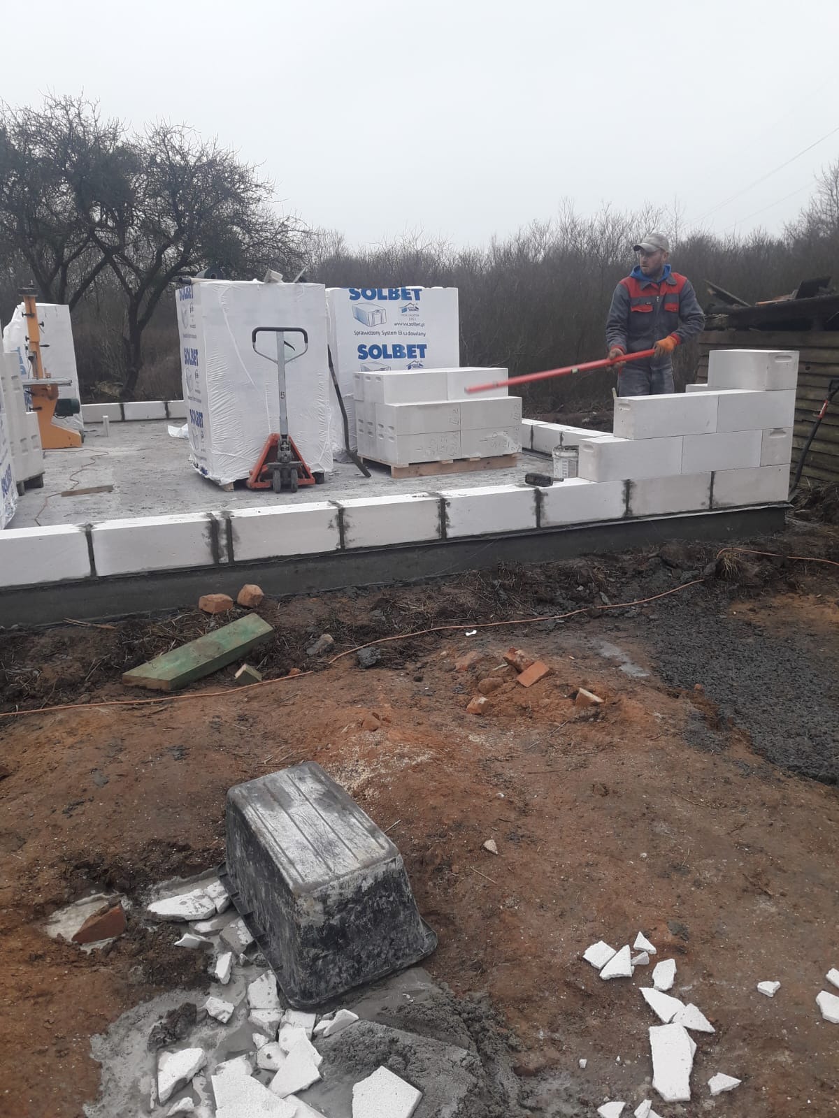 Wznoszenie ścian z białych bloczków SOLBET na fundamencie. Murarz z czerwoną pacą wyrównuje bloczki. Palety z bloczkami i wózek paletowy w tle. Grunt wokół.
