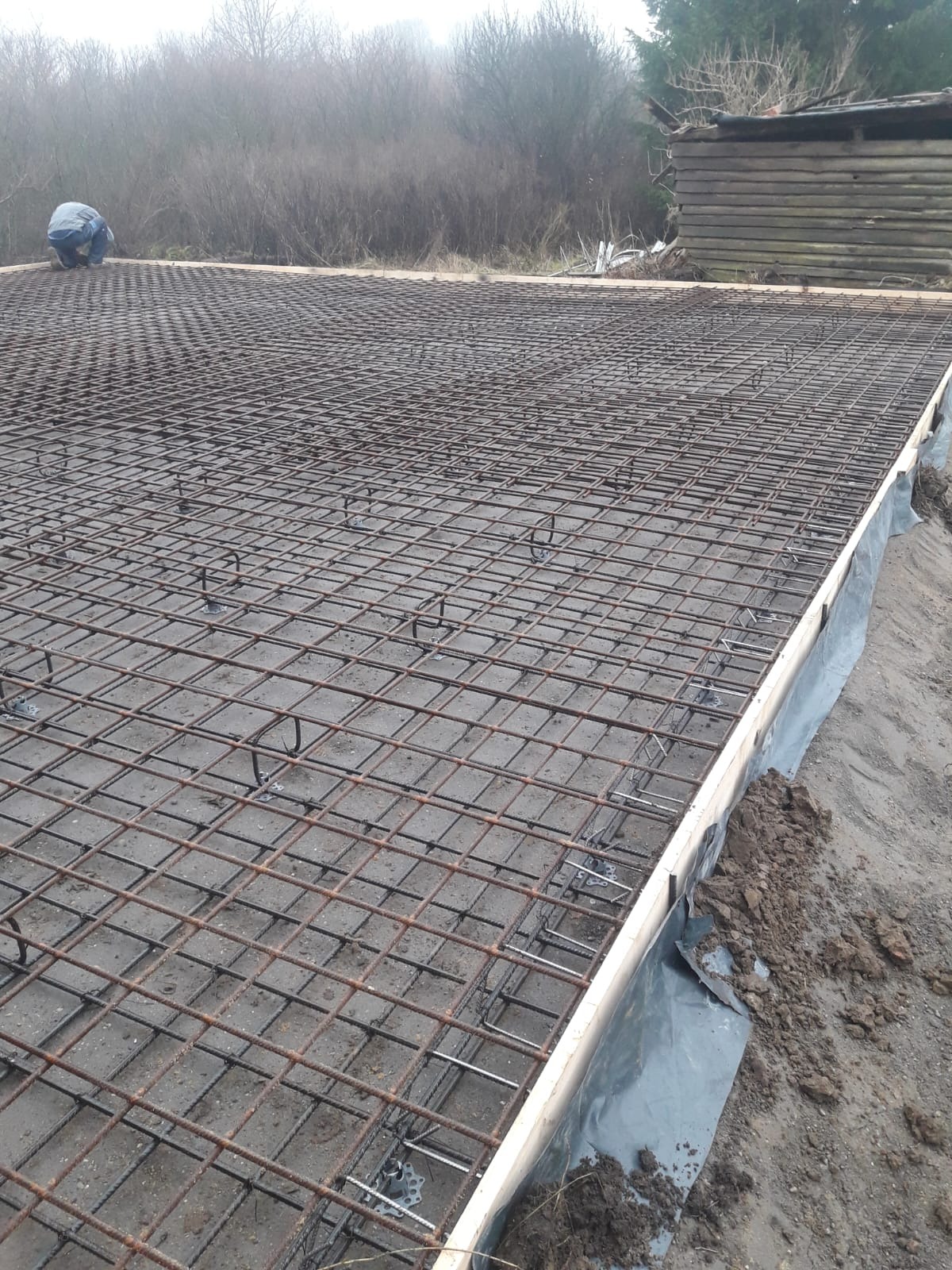 Zbrojenie fundamentu z siatki stalowej, widoczne drewniane szalunki i folia hydroizolacyjna. W tle zarys budynku gospodarczego i zarośla. Prace przygotowawcze do wylewki.