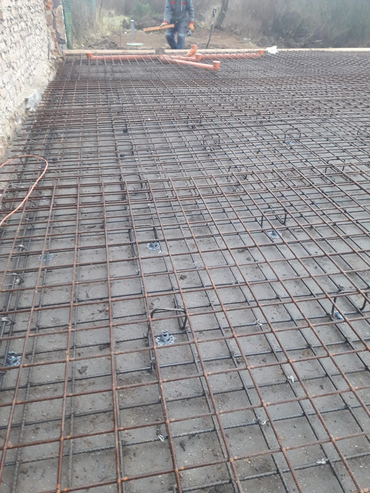 Zbrojenie fundamentów z widocznymi elementami dystansowymi i częściowo ułożonymi rurami kanalizacyjnymi. W tle pracownik budowlany.