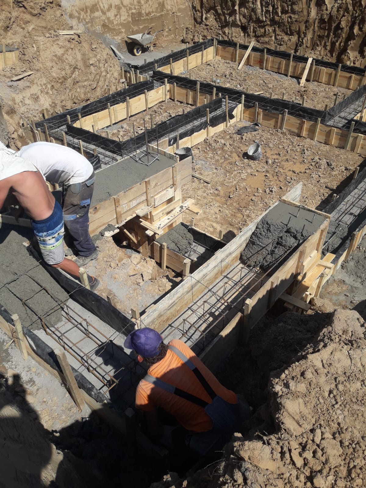 Zalewanie betonem szalunków fundamentowych zbrojonych stalą. Widoczne drewniane formy, pracownicy w trakcie wylewania betonu. Ujęcie z góry, w słoneczny dzień.