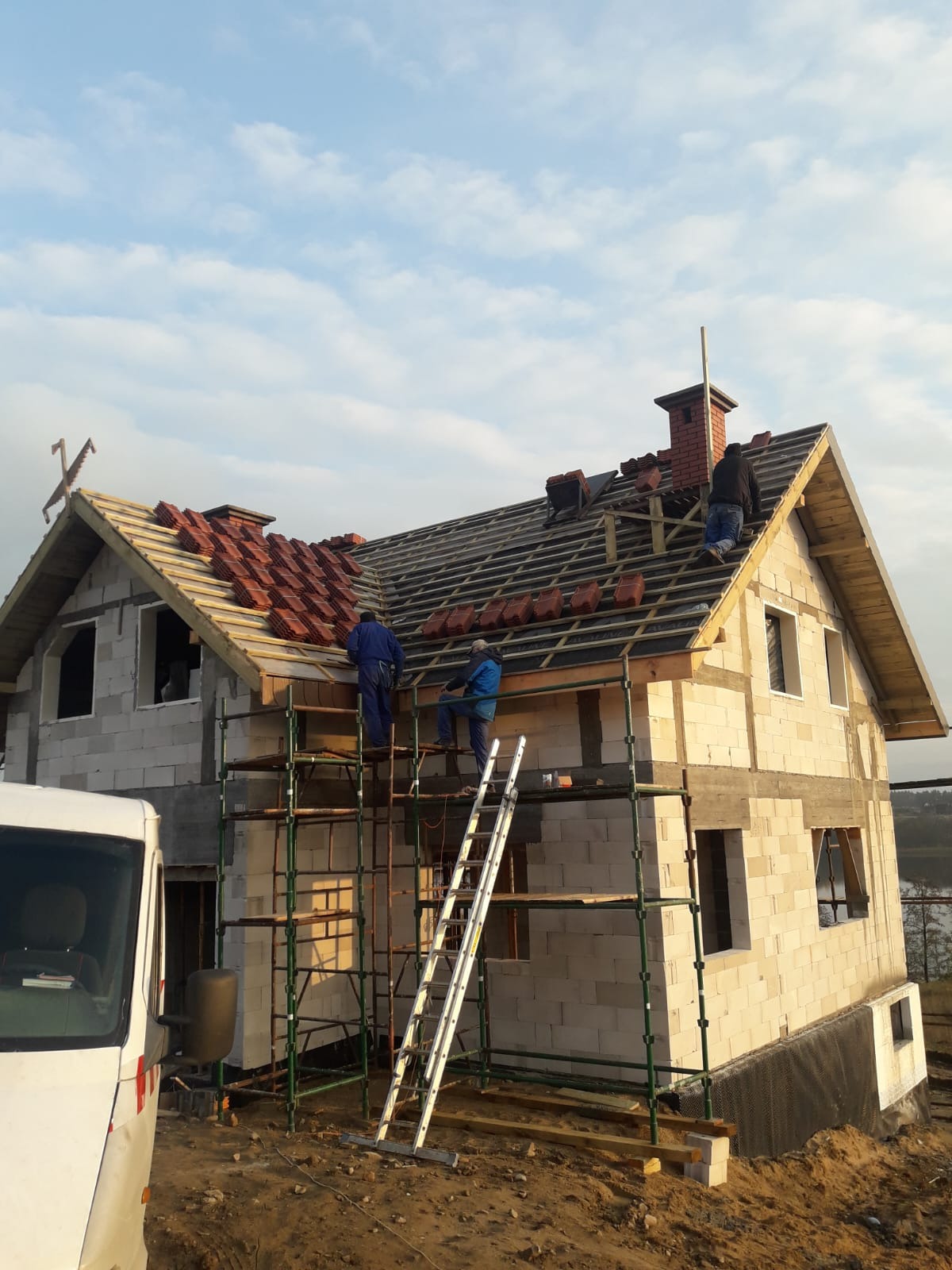 Budowa domu z białego pustaka: dekarze na rusztowaniu układają czerwoną dachówkę. Widoczny komin i fragment białego busa. Częściowo pokryty dach.