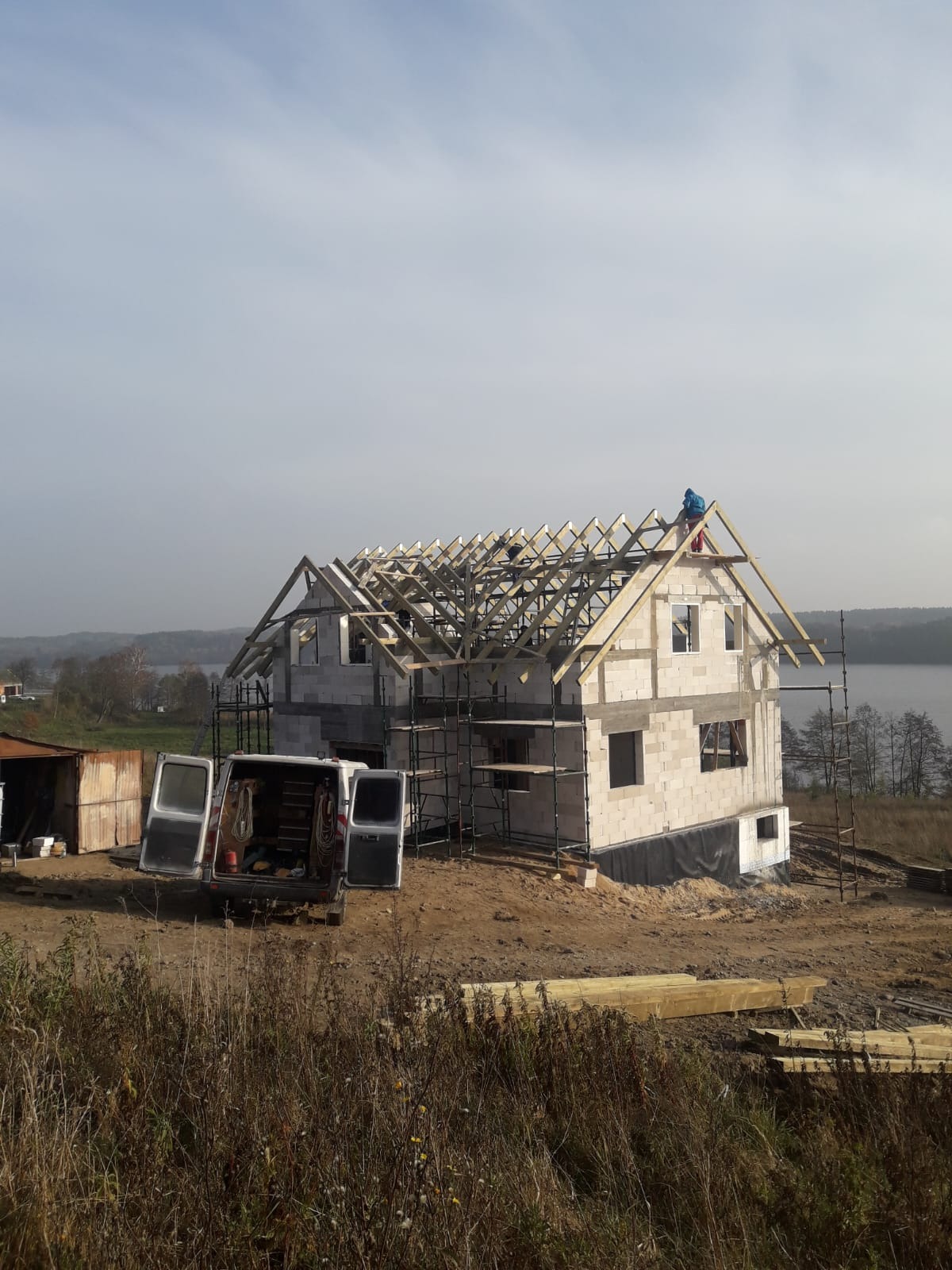 Budowa domu z białego pustaka: ściany wzniesione, więźba dachowa w trakcie montażu, rusztowania, bus z otwartymi drzwiami na tle jeziora i jesiennej roślinności.
