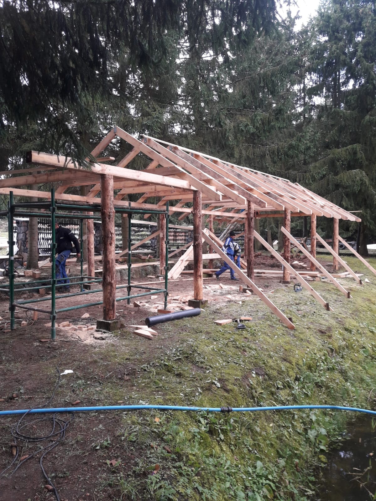 Budowa drewnianej konstrukcji z bali na zewnątrz, widoczne rusztowania i pracownicy w trakcie montażu dachu. W tle drzewa.