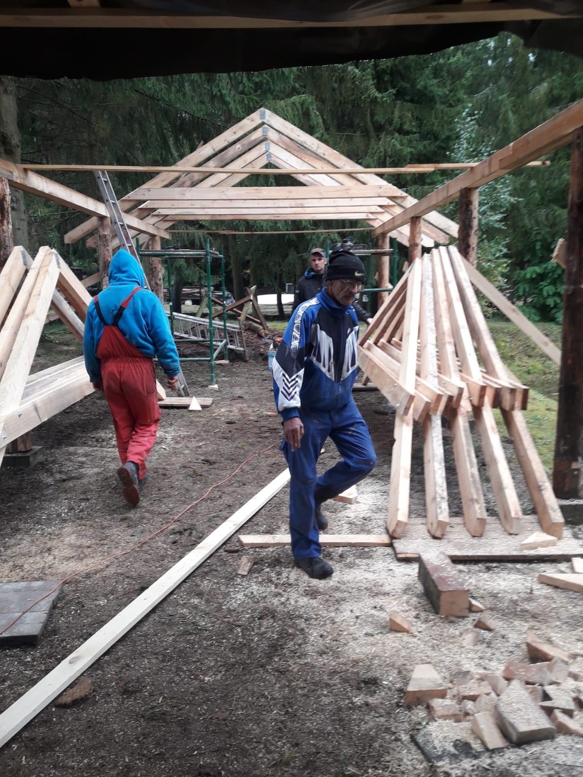 Budowa drewnianej konstrukcji dachu w lesie. Widoczni pracownicy w kombinezonach, drewniane elementy konstrukcyjne i leśne otoczenie. Prace w toku.