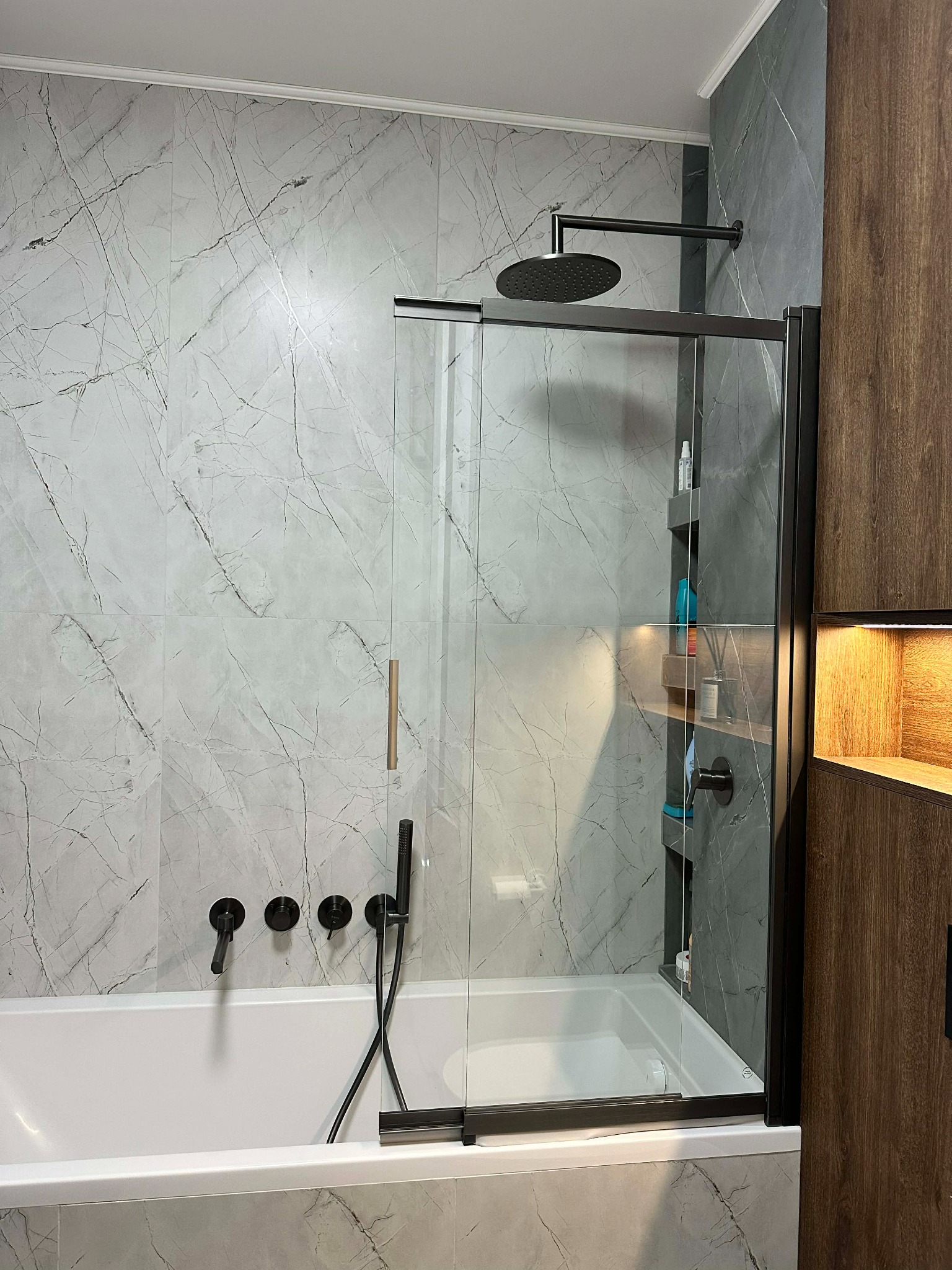 Nowoczesna łazienka z wanną, szklanym parawanem i deszczownicą. Płytki imitujące marmur w odcieniach szarości. Czarna armatura i drewniane szafki z oświetleniem.