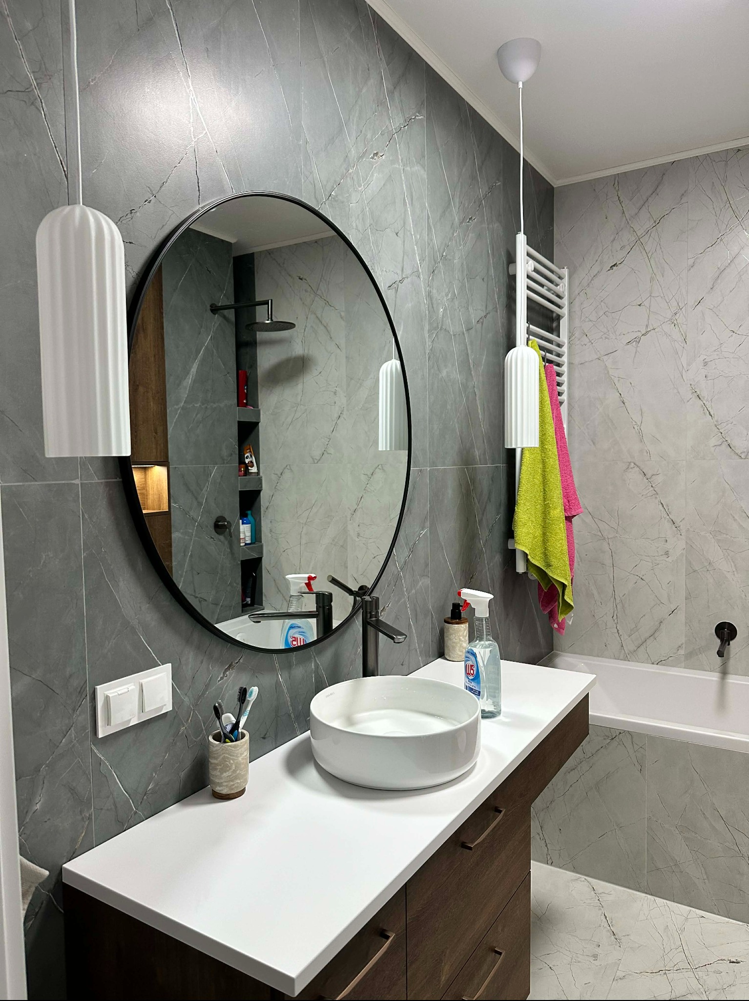 Nowoczesna łazienka z okrągłym lustrem, marmurowymi płytkami i wiszącymi lampami. Biała umywalka nablatowa i ciemne drewniane szafki pod umywalką.
