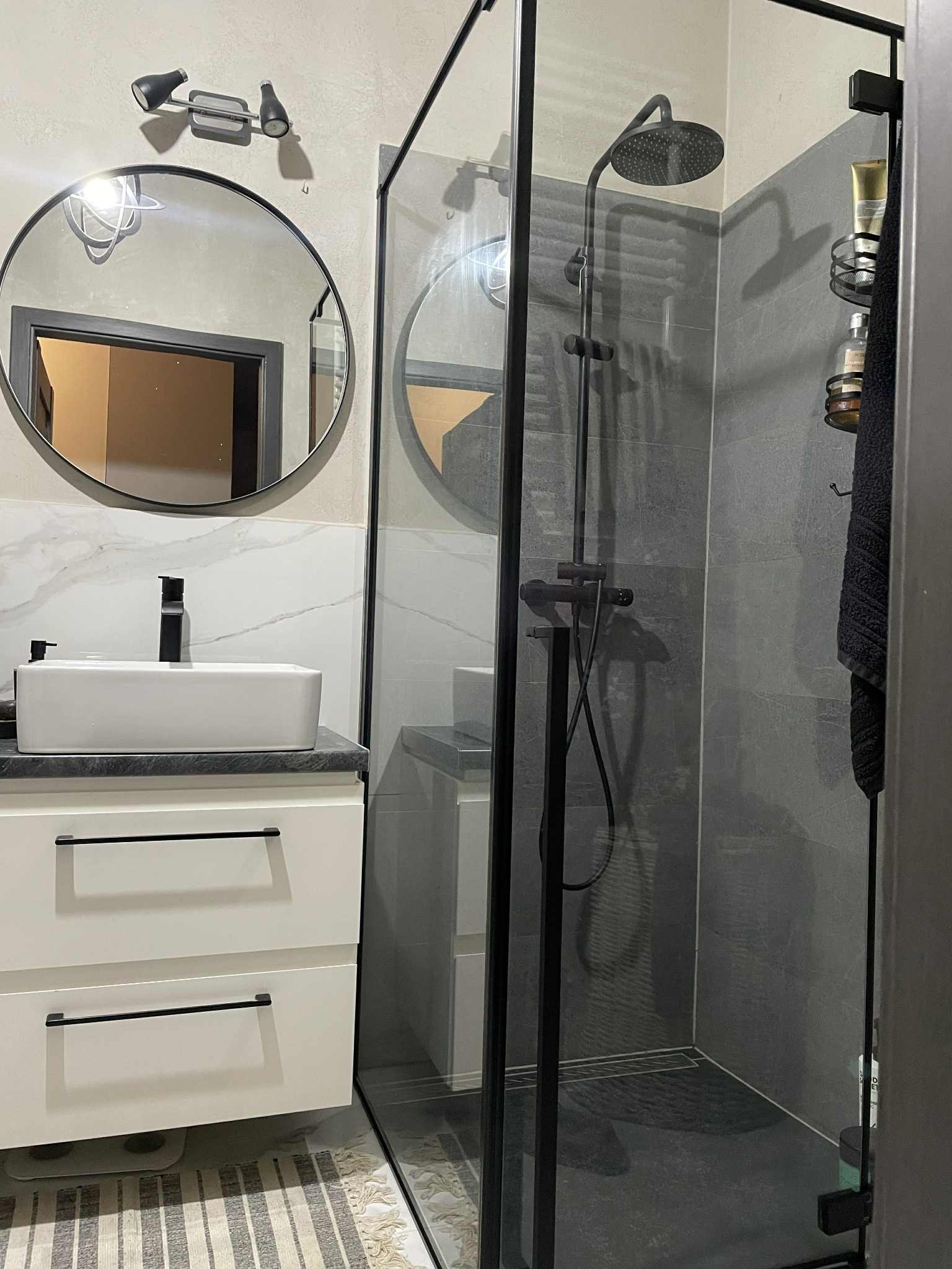 Nowoczesna łazienka z kabiną prysznicową typu walk-in, umywalką nablatową na białej szafce i okrągłym lustrem. Szare płytki i czarne akcenty tworzą elegancki wystrój.