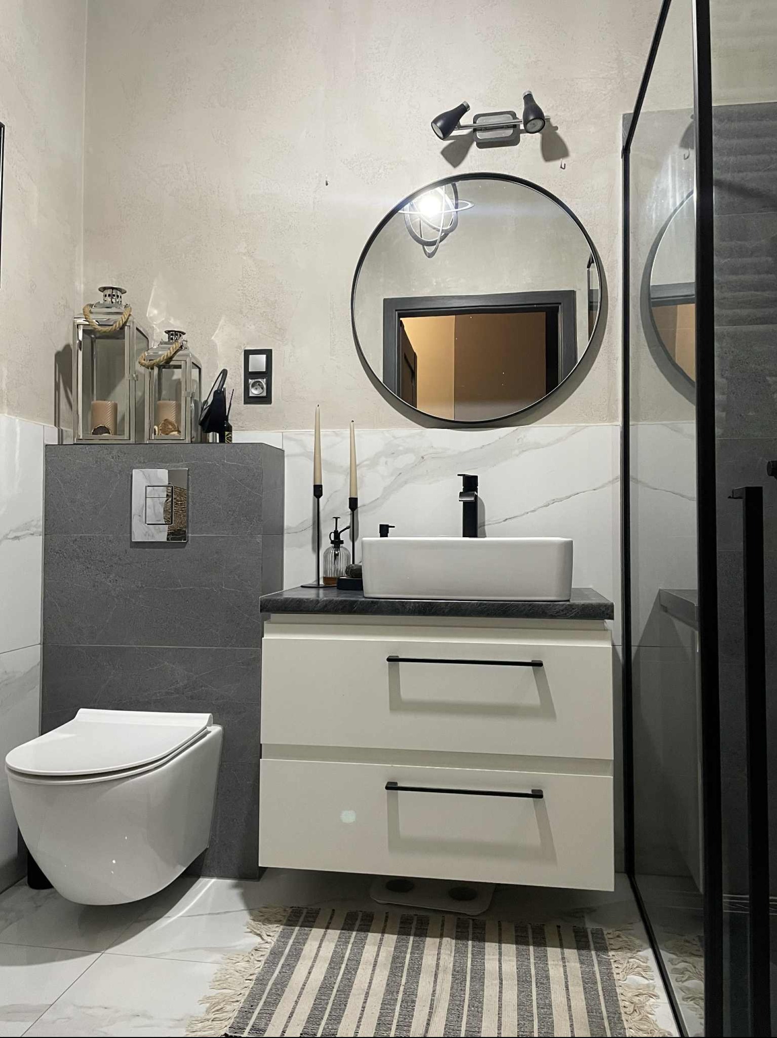 Nowoczesna łazienka z wiszącą toaletą, umywalką nablatową i okrągłym lustrem. Szaro-biała kolorystyka, minimalistyczne dodatki, kabina prysznicowa z czarnymi profilami.