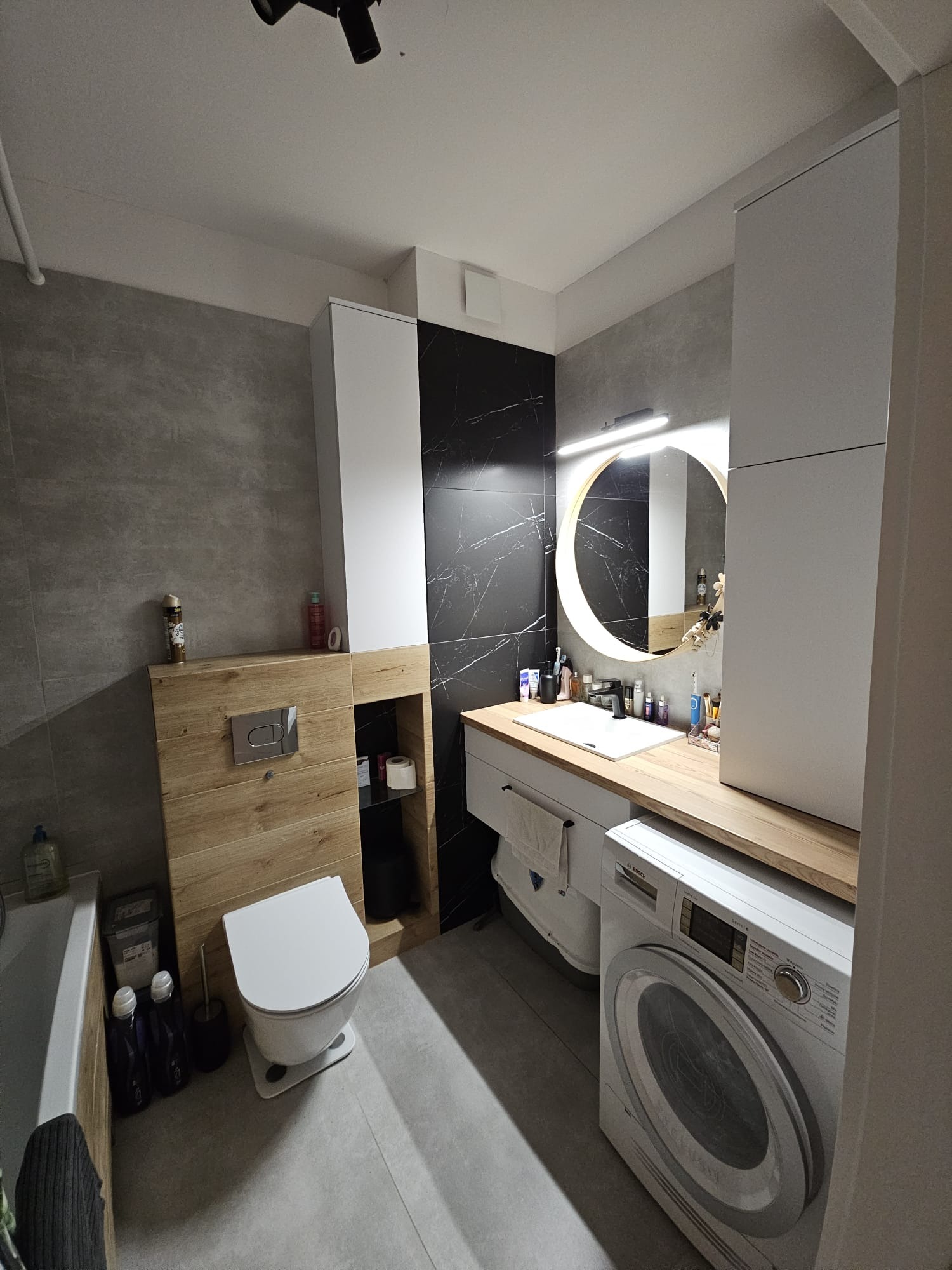 Nowoczesna łazienka z drewnianymi i marmurowymi akcentami. Okrągłe lustro z podświetleniem, biała umywalka i pralka. Minimalistyczny design, szare płytki.