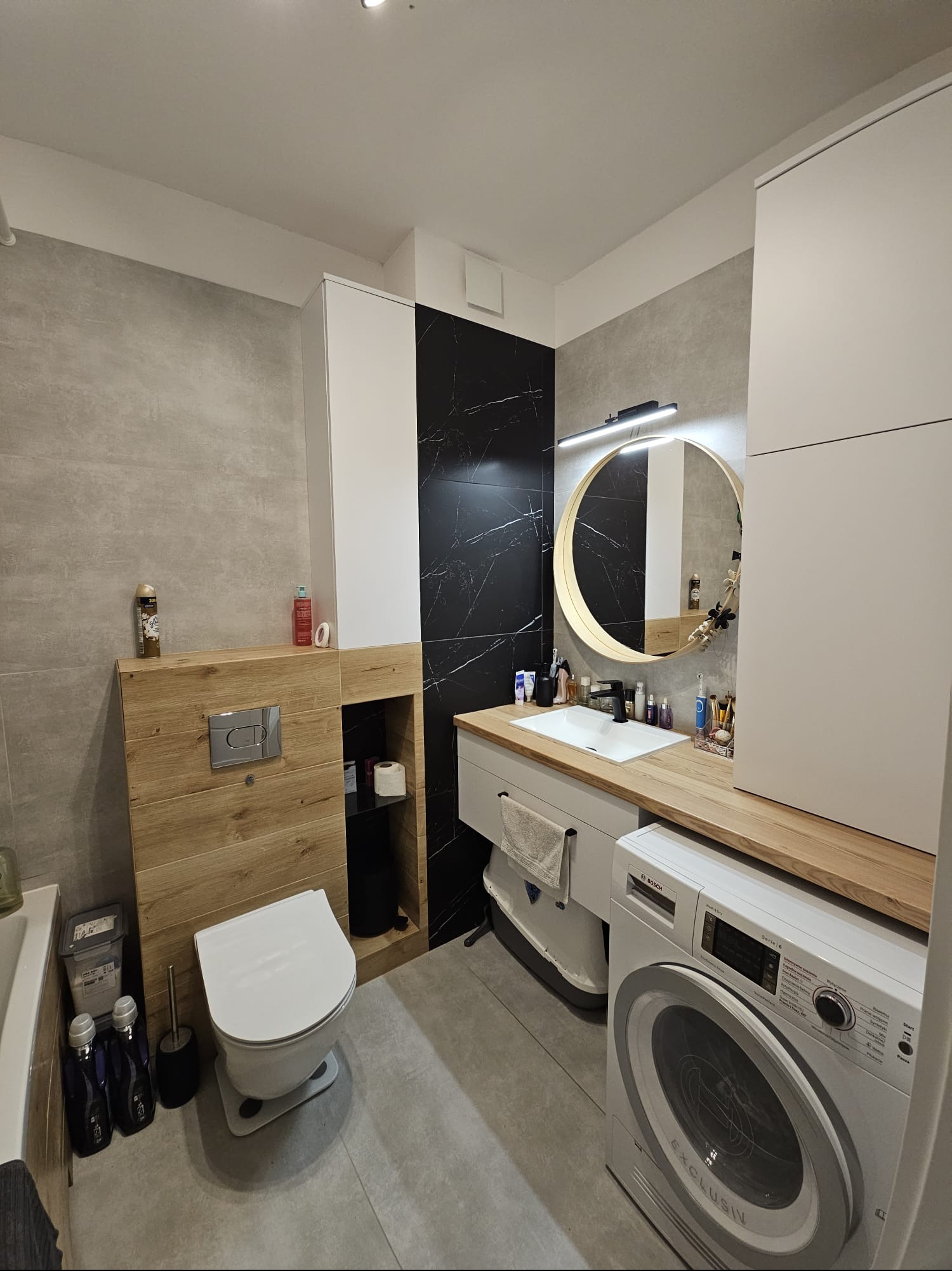 Nowoczesna łazienka z szarą płytką, czarnym marmurem i drewnianymi akcentami. Okrągłe lustro z podświetleniem nad umywalką, pralka Bosch. Minimalistyczny design.
