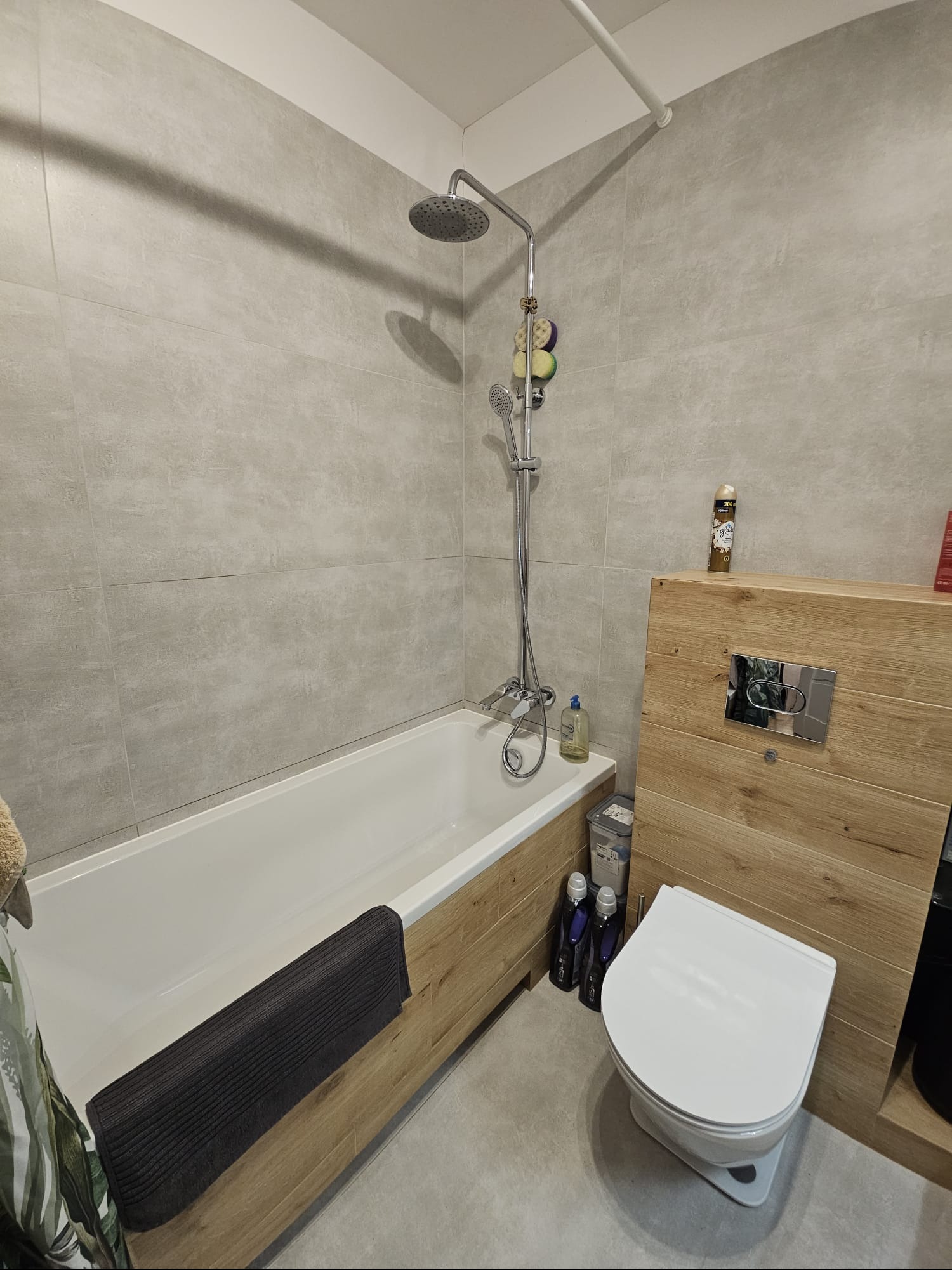 Nowoczesna łazienka z wanną obudowaną drewnem, szarą armaturą i płytkami w odcieniach szarości. Uwagę zwraca drewniana zabudowa toalety.