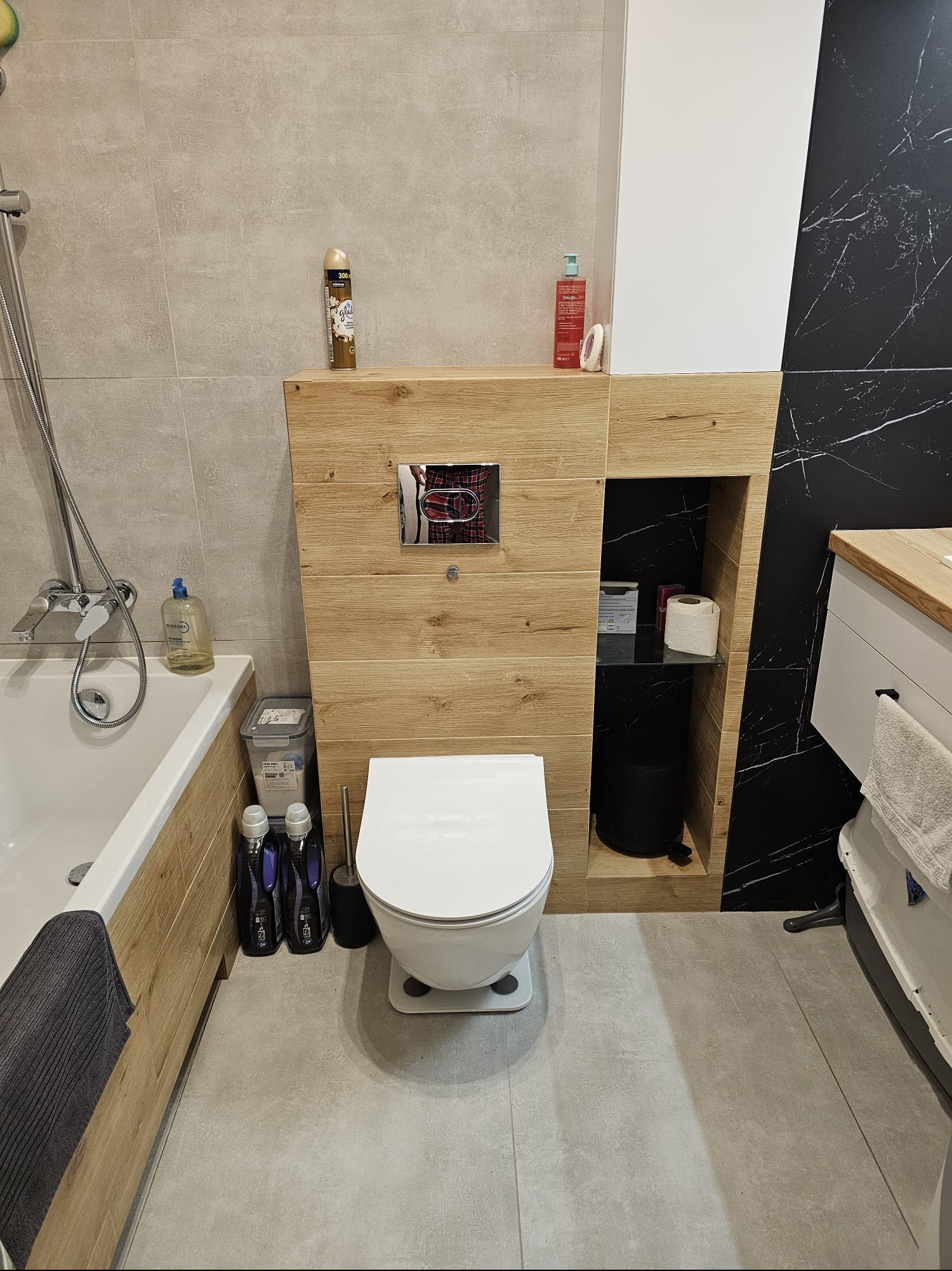 Nowoczesna toaleta z zabudową z drewnopodobnych paneli, czarnym marmurem i szarą podłogą. Widoczne akcesoria łazienkowe i armatura. Minimalistyczny design.