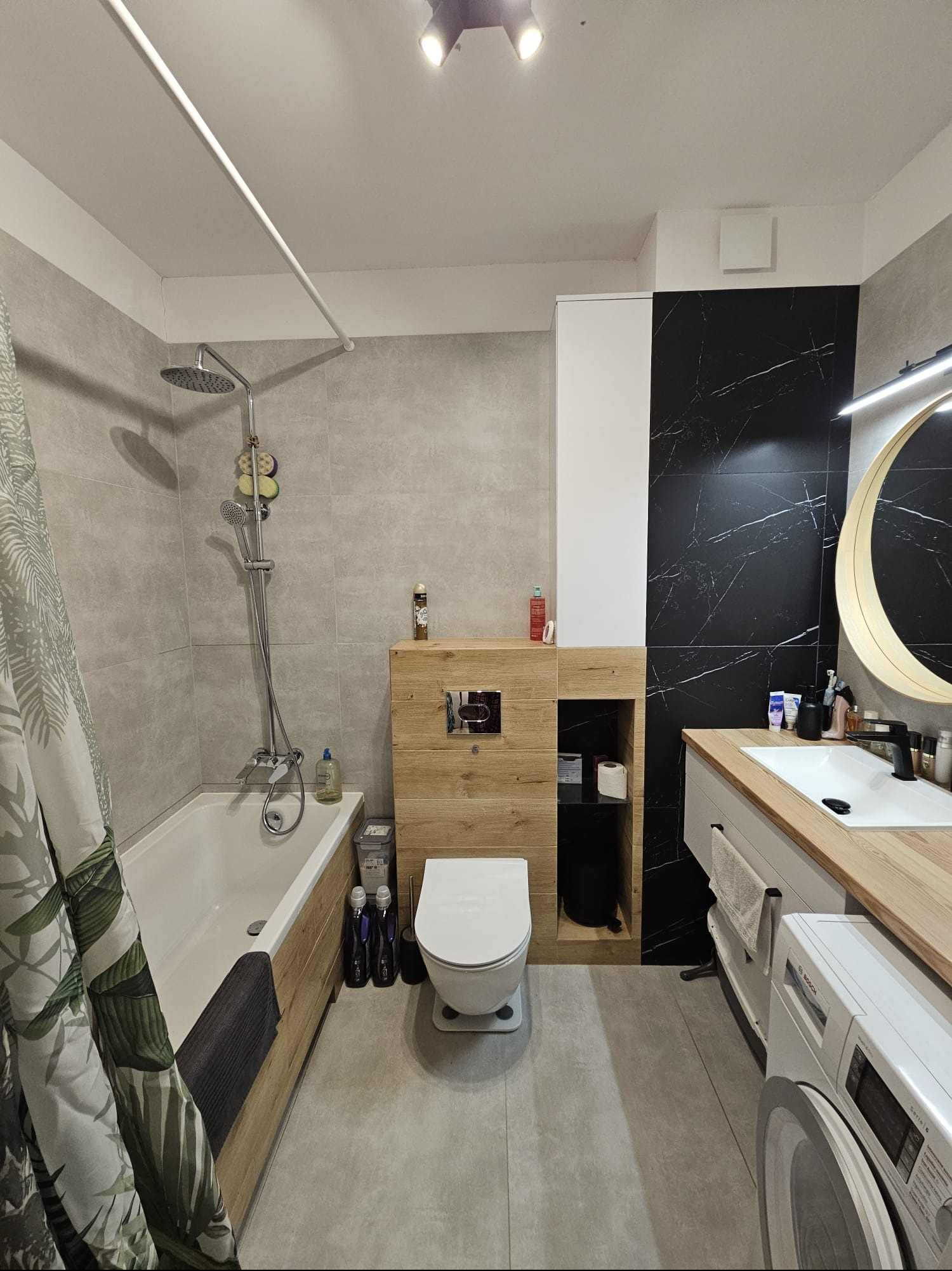 Nowoczesna łazienka z wanną, toaletą, umywalką z drewnianym blatem i pralką. Czarno-białe płytki na ścianie, okrągłe lustro z podświetleniem. Minimalistyczny design.