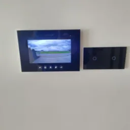 Czarny panel wideodomofonu Vidos zamontowany na białej ścianie, obok szklany panel z dotykowymi włącznikami światła.