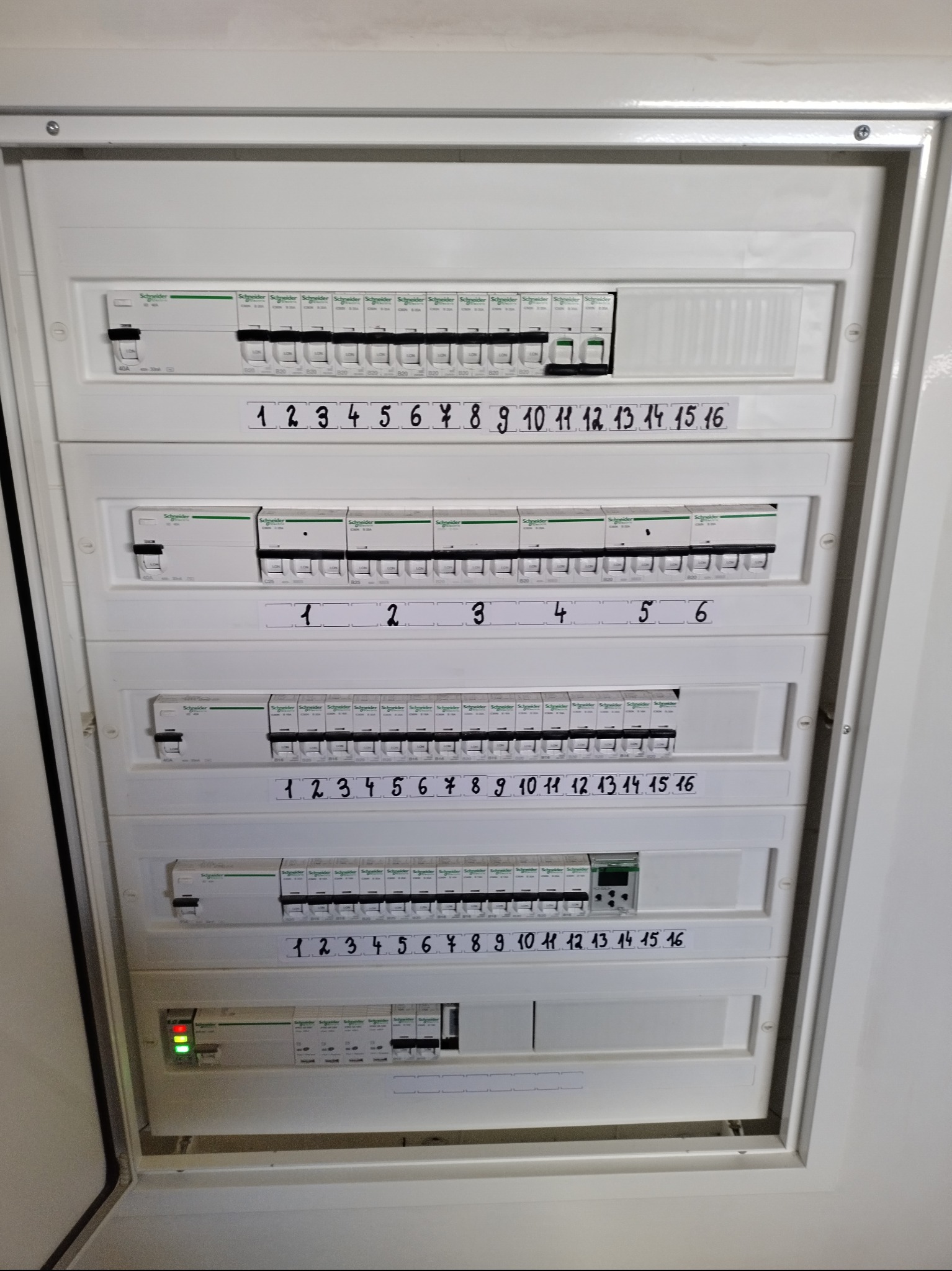 Rozdzielnica elektryczna marki Schneider z ponumerowanymi bezpiecznikami, widoczne naklejki z numerami 1-16 i 1-6, zielona dioda sygnalizacyjna po lewej stronie