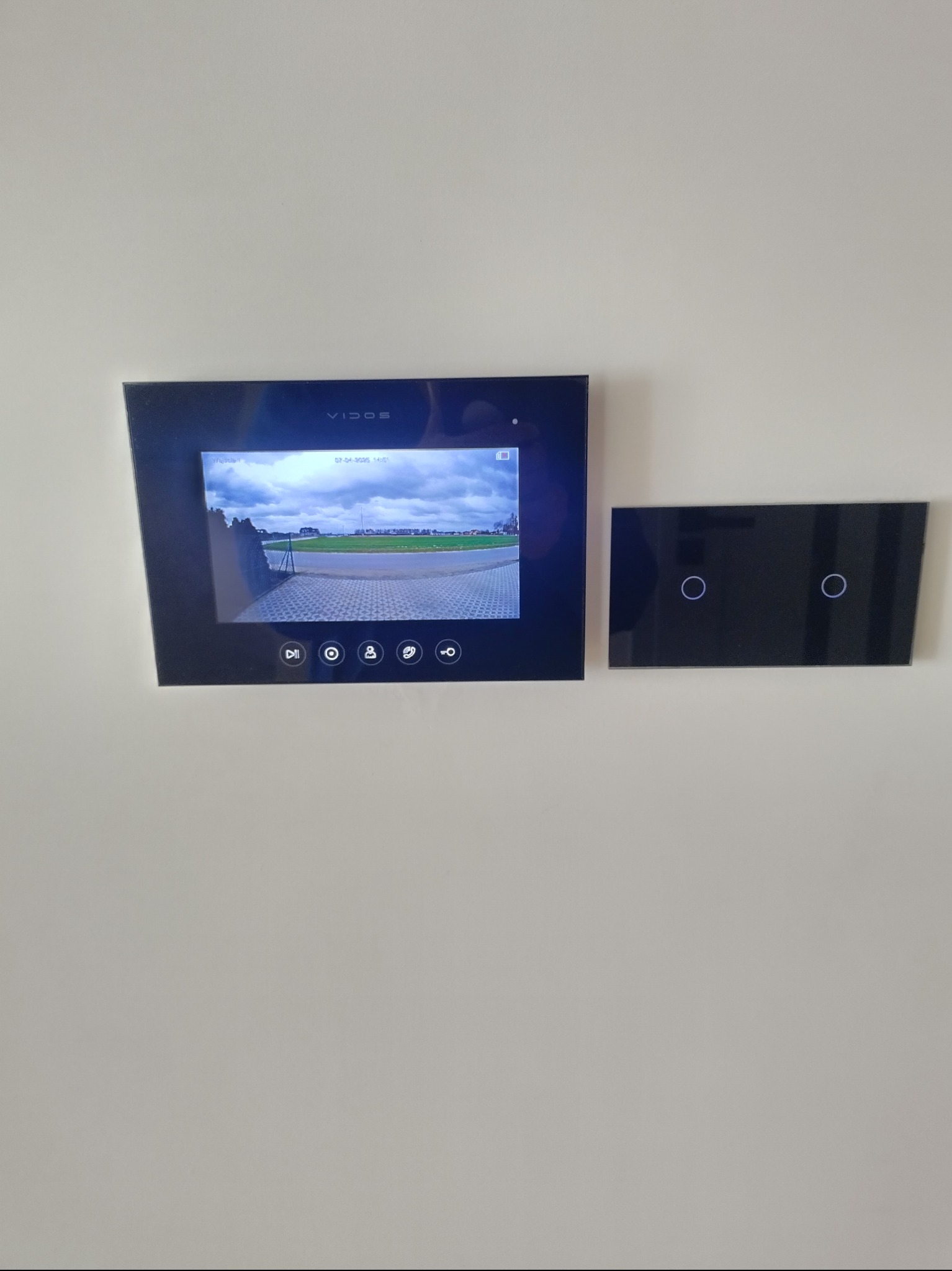 Czarny panel wideodomofonu Vidos zamontowany na białej ścianie, obok szklany panel z dotykowymi włącznikami światła.