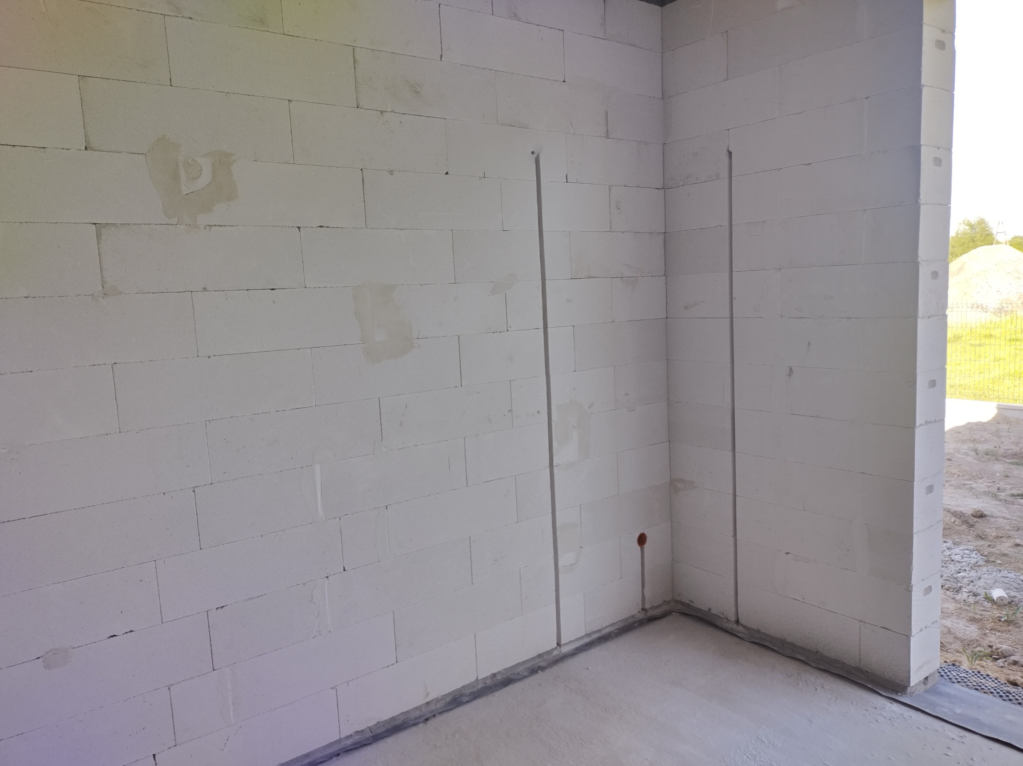Wewnętrzny róg ściany z białych bloczków, z wyżłobieniami pionowymi pod instalację elektryczną i otworem na puszkę, widok na zewnątrz przez otwór okienny.