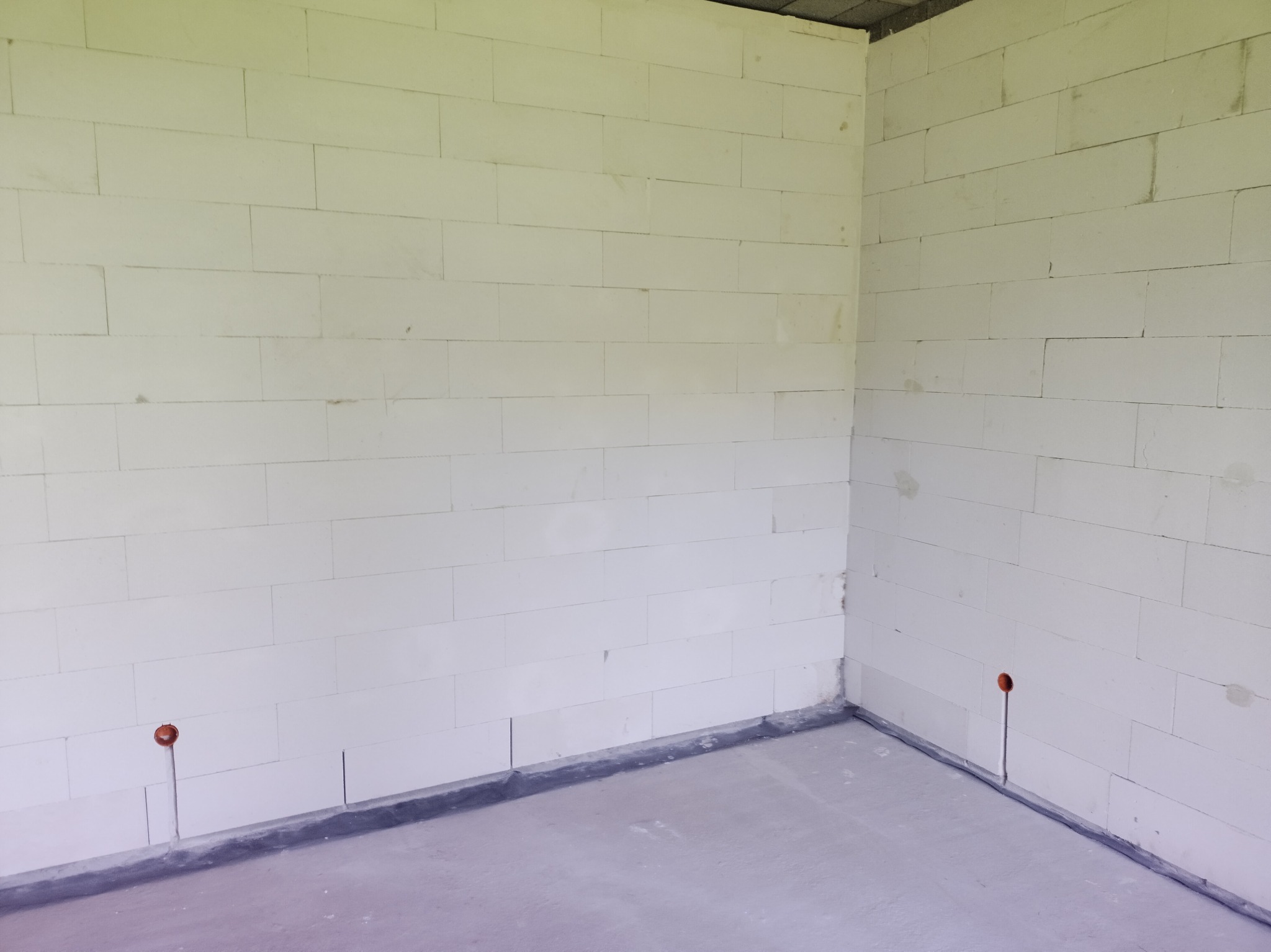 Surowy stan wnętrza: ściany z białych bloczków, w których widoczne są pionowe bruzdy z wystającymi przewodami elektrycznymi zakończonymi puszkami, szara wylewka podłogowa.