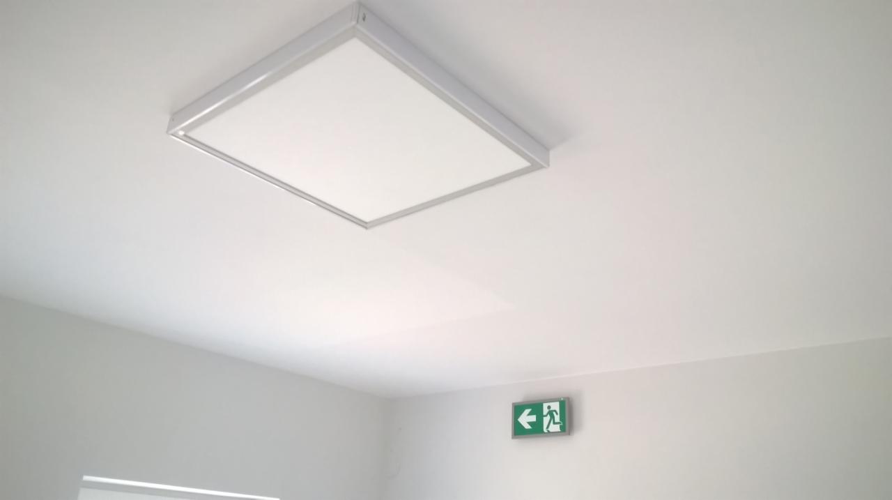Kwadratowa oprawa oświetleniowa LED zamontowana na białym suficie, obok zielony znak ewakuacyjny ze strzałką w lewo.