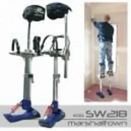 Szczudła malarskie marki Marshalltown SW218 stojące obok pracującego malarza na szczudłach, malującego ścianę w narożniku pokoju, widoczne ślady po taśmie malarskiej na podłodze.