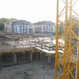 Budowa wielorodzinnego budynku mieszkalnego w trakcie wznoszenia fundamentów, widoczny szkielet zbrojeniowy, szalunki i żółty żuraw wieżowy na pierwszym planie, w tle gotowe budynki.