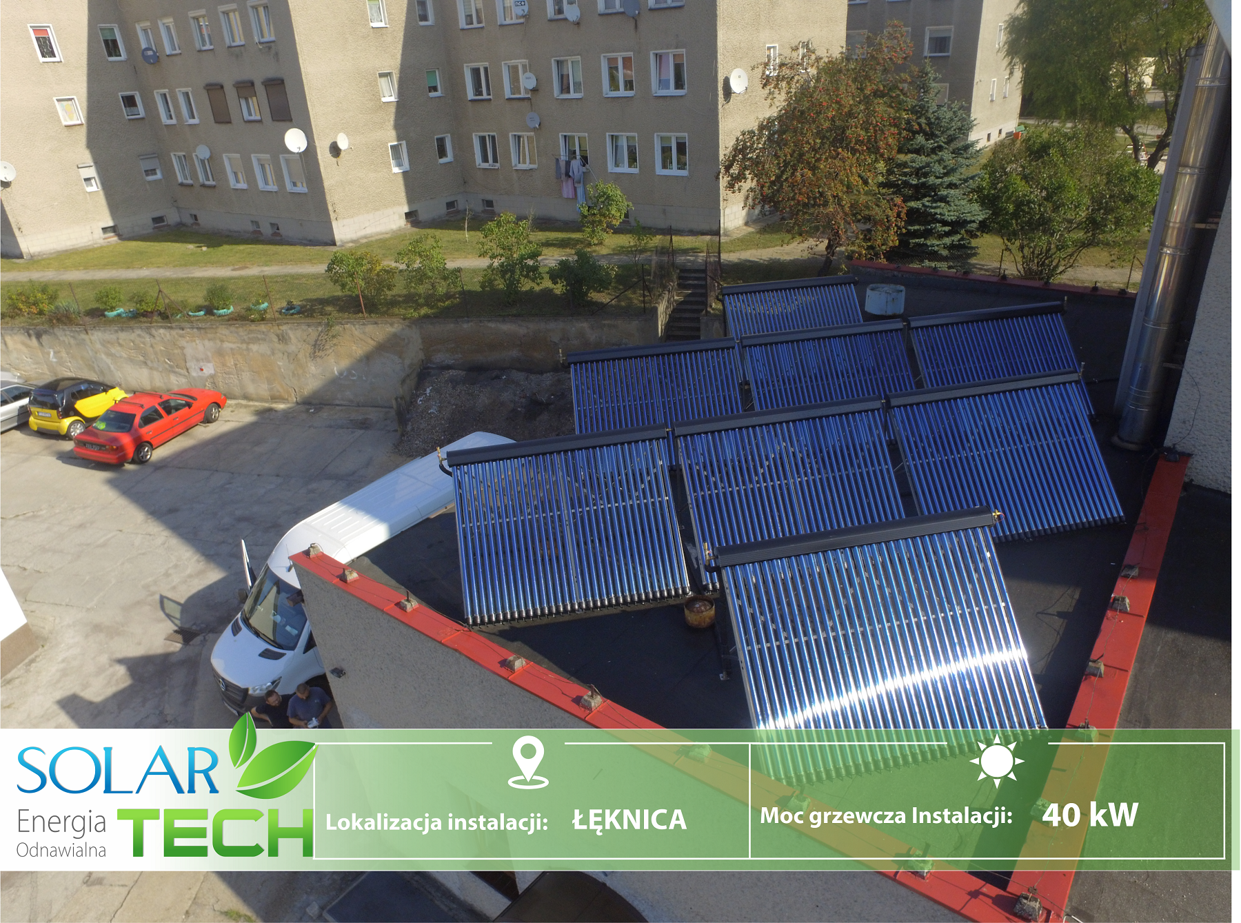 Widok z góry na instalację kolektorów słonecznych na dachu budynku mieszkalnego, z widocznym budynkiem w tle, zaparkowanymi samochodami i logotypem firmy SOLAR-TECH.