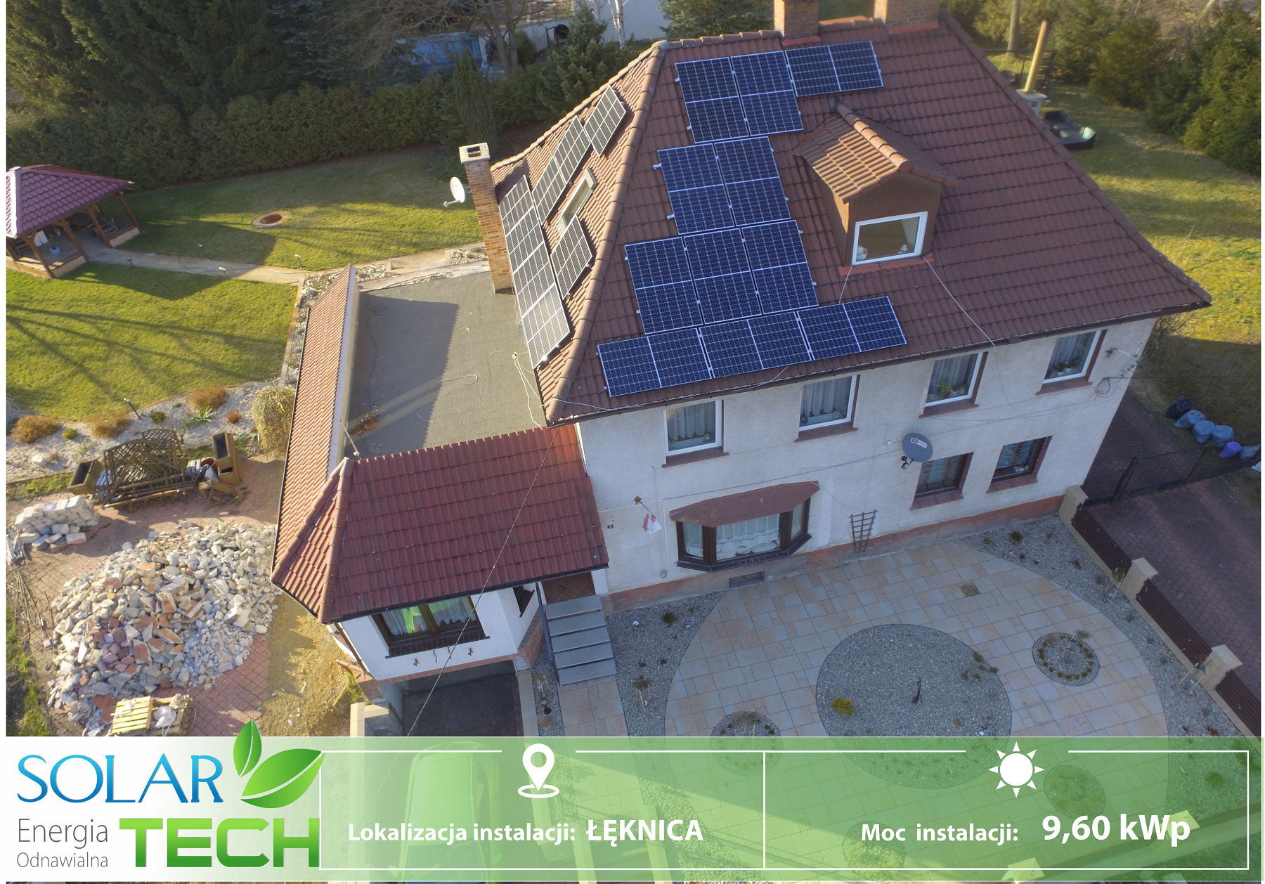 Widok z góry na dom z panelami słonecznymi na dachu, otoczony zielenią i brukowanym podjazdem. W tle widoczny ogród z altaną i stertą gruzu.