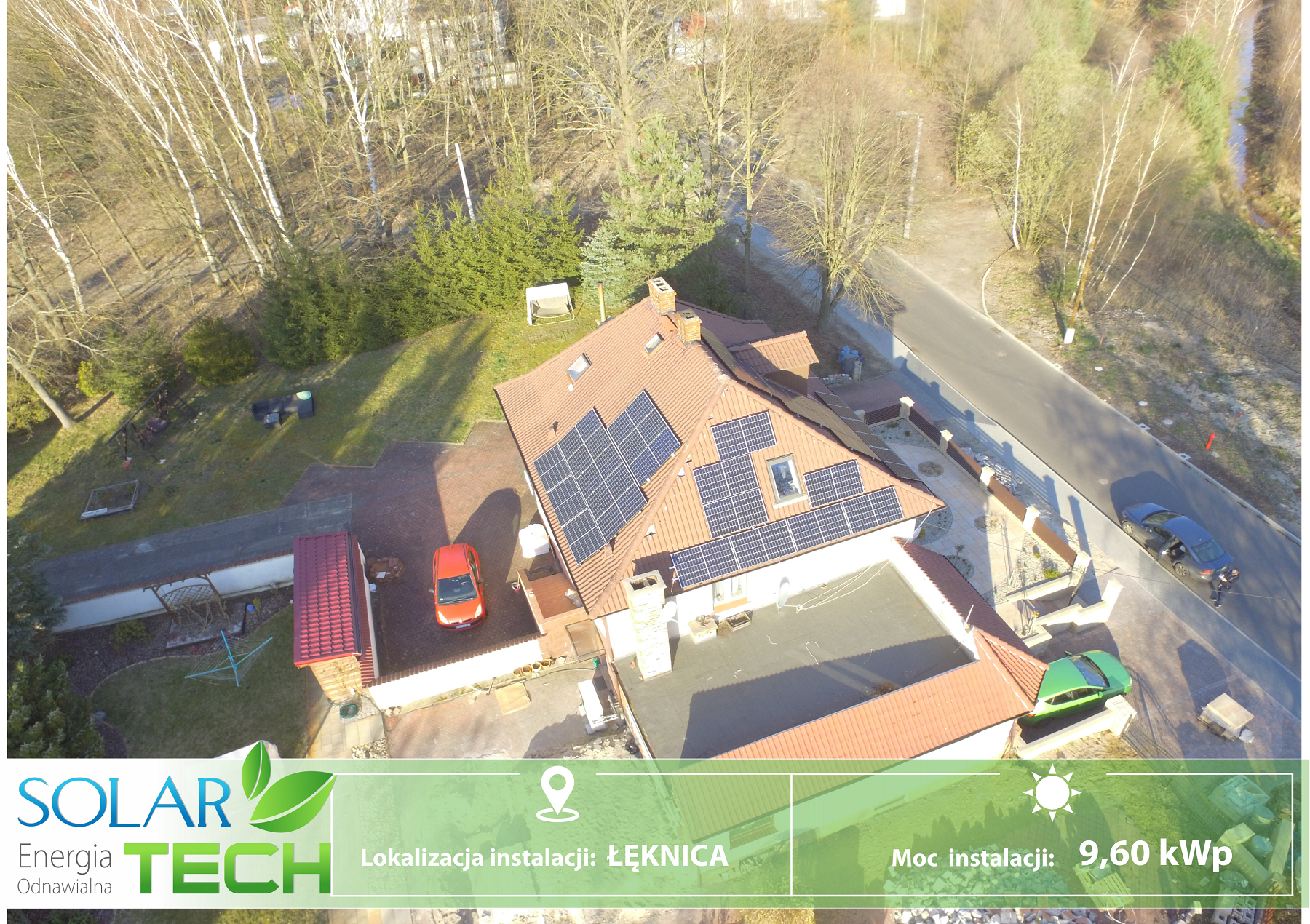 Widok z lotu ptaka na dom z panelami słonecznymi na dachu, czerwony samochód zaparkowany obok, otoczony drzewami i drogą.