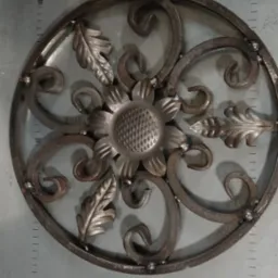 Ozdobny, metalowy element dekoracyjny w kształcie koła z motywem kwiatowym i liśćmi, połączony spawami punktowymi, leżący na metalowym stole z podziałką.