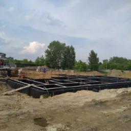 Przygotowanie fundamentów pod budynek: wykop zabezpieczony czarną folią, zbrojenie stalowe, pracownicy budowlani i koparka na placu budowy w słoneczny dzień.