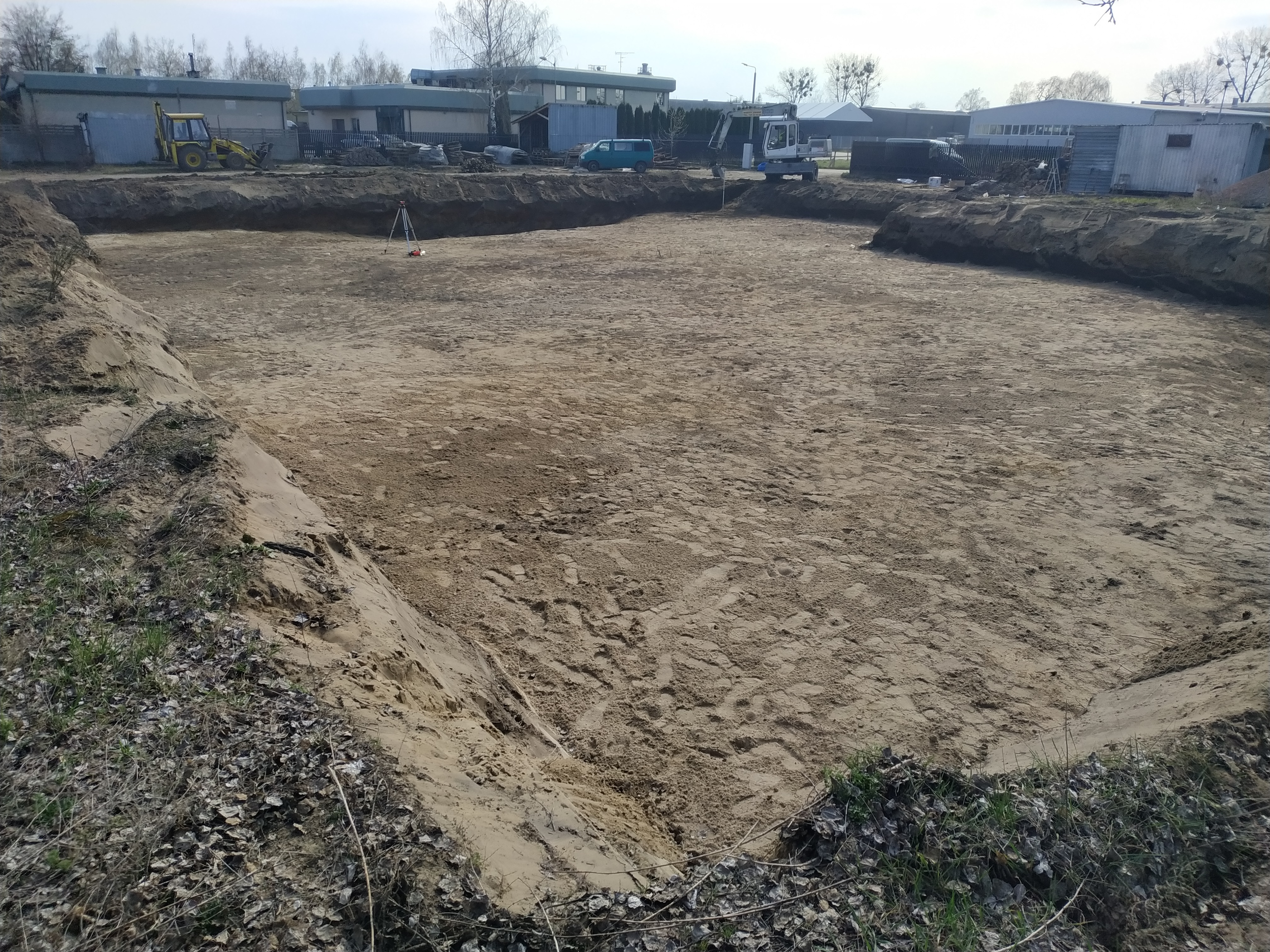 Duży, prostokątny wykop w piaszczystej ziemi, z widocznymi śladami maszyn i narzędzi pomiarowych; w tle budynki gospodarcze i sprzęt budowlany.