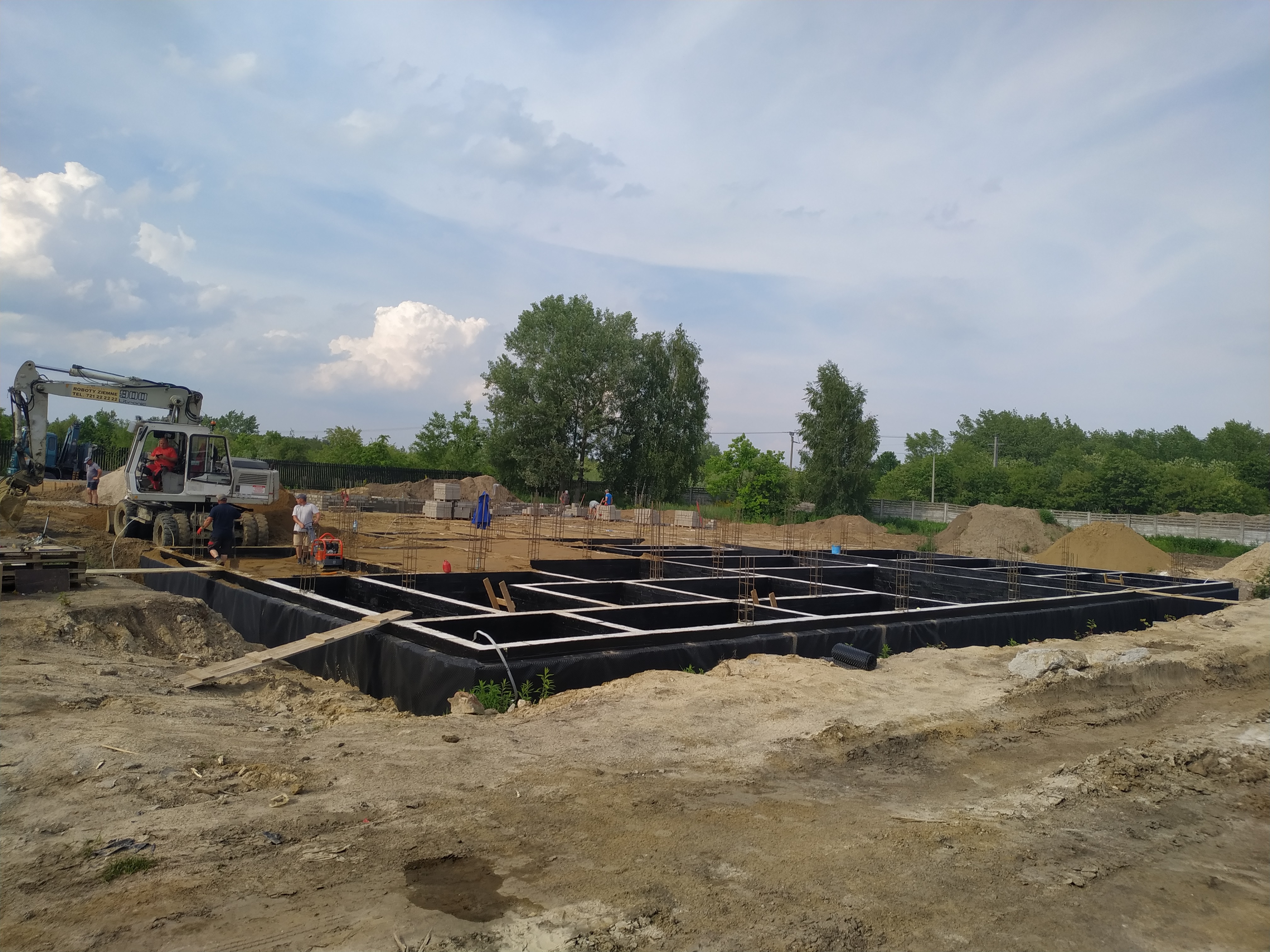 Przygotowanie fundamentów pod budynek: wykop zabezpieczony czarną folią, zbrojenie stalowe, pracownicy budowlani i koparka na placu budowy w słoneczny dzień.